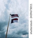 Two flags waving in the wind. One American flag and the other is a North Carolina flag. Flag pole with rope. Background is dramatic with dark rain clouds.