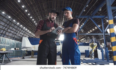 Two flight engineers walking through a large aircraft hangar talking and gesturing together - Powered by Shutterstock