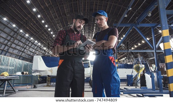 Two flight engineers walking\
through a large aircraft hangar talking and gesturing\
together