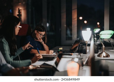 Two focused female colleagues collaborate at a late-night business meeting in a well-lit office setting, indicating teamwork and dedication. - Powered by Shutterstock