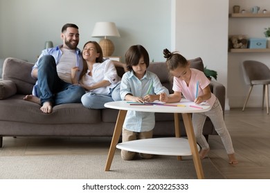 Two focused little sibling kids playing games at home, drawing in colored pencils in living room, enjoying learning creative activity. Happy young couple of parents relaxing on sofa in background - Shutterstock ID 2203225333