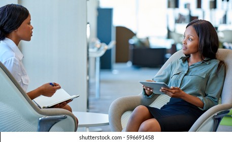 Two Focused Young Business Colleagues Sitting In Chairs Talking Together While Having A Meeting In A Modern Office