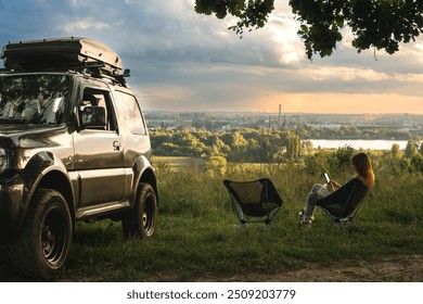 Two folding light weight, portable chairs and woman reading the book. Nature, on the top, river, sunset. Tourism and active lifestyle. Equipment for tourism. Off road vehicle with roof rack - Powered by Shutterstock