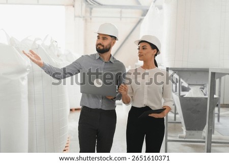 Two food processing engineers wearing white hardhats and holding laptop and tablet inspect large sacks of flour in a food processing plant
