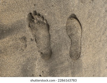 Two footmarks side by side: one imprint of bare human foot and another print of shoe in beach sand in Lanzarote, Canary islands. Footsteps of bare and shod feet on beach - different purposes of people