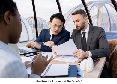 Two Foreign Intercultural Business Partners Discussing Terms Of Contract At Meeting While Sitting By Table In Cafe