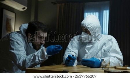 Two forensic experts in protective suits examine and discuss evidence under artificial light, suggesting a late-night crime investigation.