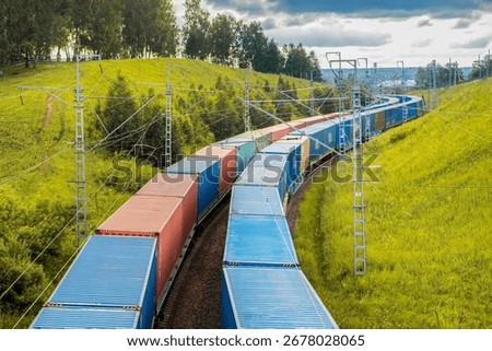 Two freight trains move in parallel, carrying colorful containers along railway tracks. Set against a backdrop of vibrant green hills, the natural beauty enhances the scene as the trains