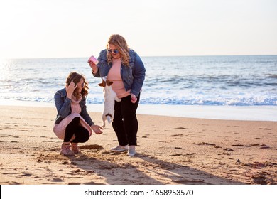 Two Freinds And Dog Walking On The Spring Beach. Fat Woman With Strong Friend Playing With Pet. Overweight Woman Dressed Jeans Jacket And Pink Sweater. People Enjoing The Nature And Evening Sun