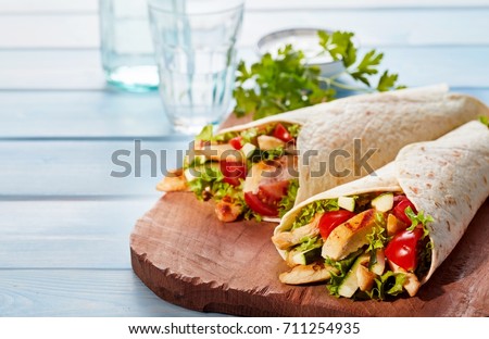 Two fresh chicken and salad tortilla wraps on wooden cutting board with glasses in background