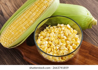 Two fresh ears of corn, one partially husked, next to a bowl of sweet corn kernels, all arranged on a wooden cutting board.