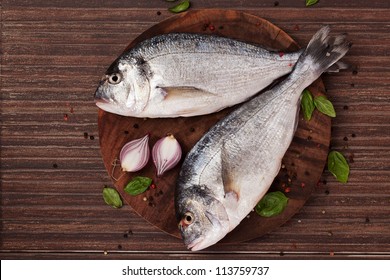 Two fresh gilt hear sea bream on round wooden chopping board with fresh basil leaves, colorful peppercorn and onion, top view. Seafood coocking ingredients in natural brown.