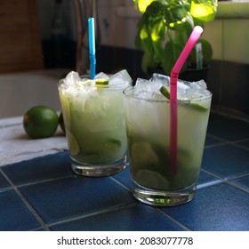 Two Fresh Iced Caipirinhas On A Blue Tiled Counter Top With Blue And Pink Straws. A Lime And Basil Plant Sit In The Background.