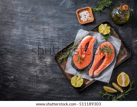 Two fresh raw salmon steaks on paper with salt, peppers, lemon, and rosemary on black background, top view