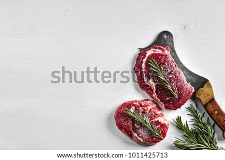 Two fresh raw steaks with herbs, garlic, olive oil, pepper, salt, rosemary and a kitchen ax on white background. Top view.