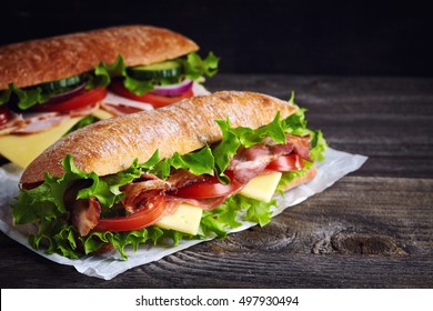 Two fresh submarine sandwiches with ham, cheese, bacon, tomatoes, lettuce, cucumbers and onions on dark wooden background - Powered by Shutterstock