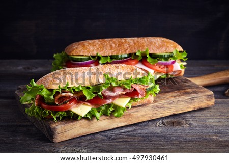 Two fresh submarine sandwiches with ham, cheese, bacon, tomatoes, lettuce, cucumbers and onions on wooden cutting board