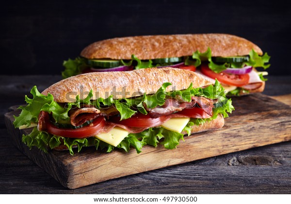 Two\
fresh submarine sandwiches with ham, cheese, bacon, tomatoes,\
lettuce, cucumbers and onions on wooden cutting\
board