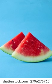Two fresh triangular watermelon wedges with green rind standing together on a bright blue solid background, vertical composition.