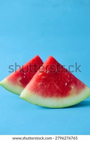 Two fresh triangular watermelon wedges with green rind standing together on a bright blue solid background, vertical composition.