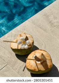Two fresh young green coconuts with straws, decorated with a white plumeria flower on a stone deck next to a blue swimming pool.