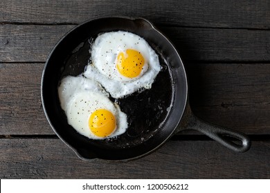 Two fried eggs in cast iron frying pan sprinkled with ground black pepper. Isolated on dark painted wood from above.