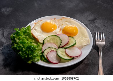 Two fried eggs with a leaf of lettuce, cucumber and radish slices on a white plate. Scrambled eggs. Breakfast. Healthy eating. Close-up