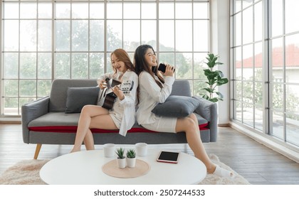 Two friend sit on sofa in a bright living room with large glass wall. Play guitar and use smartphone as microphone, sing happily. Tablet computer, coffee cup and decorated pot plant are on the table.
 - Powered by Shutterstock