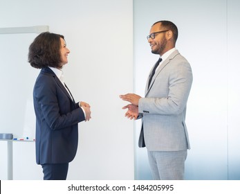 Two Friendly Business Colleagues Discussing Training In Conference Room. Business Man And Woman Standing Indoors Near White Board, Talking And Laughing. Business Colleagues Concept