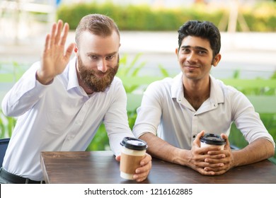 Two Friendly Business Men Drinking Coffee In Outdoor Cafe. One Man Waving His Hand To Viewer With Blurred Plants In Background. Coffee Break Concept. Front View.