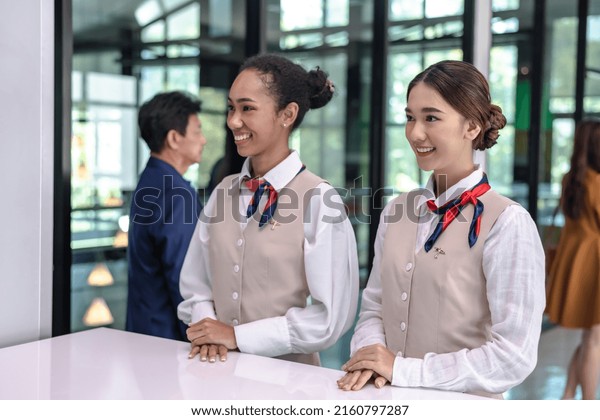 Two friendly female airport ground staffs in\
airline uniforms standing at airport check in counter,airline\
workers working at airport,smiling airline ground workers at check\
in desk, aviation business