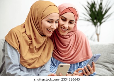 Two friendly muslim women look at the phone and rejoicing at good news or new purchases. Girls holding a phone in their hands Sitting on the sofa in a homely atmosphere. Beautiful arabs read messages - Powered by Shutterstock