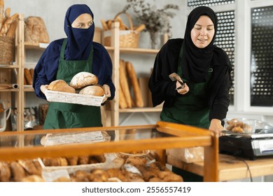 Two friendly Muslim women in traditional clothes working in cozy local bakery, arranging freshly baked bread on shelves and weighing pastries for customer with welcoming smile - Powered by Shutterstock