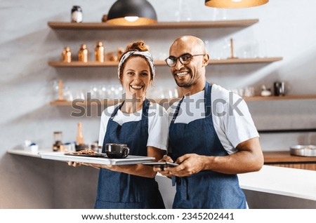 Two friendly waiters stand together in a coffeehouse, holding a tray of food and tablet as they get ready to provide excellent service to customers. Professional waitstaff working as a team.