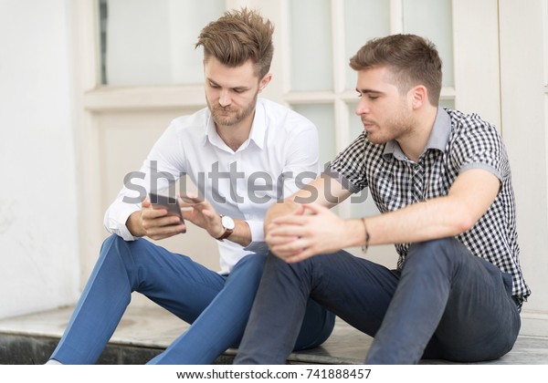 Two friends in casual wear uses\
smartphone sitting together. Best friends enjoying\
concept.