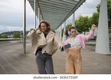 Two friends dancing joyfully while sharing music through headphones and a smartphone in an urban setting, embracing their free time and enjoying each other's company - Powered by Shutterstock - Get 15% off with code: PIKWIZARD15