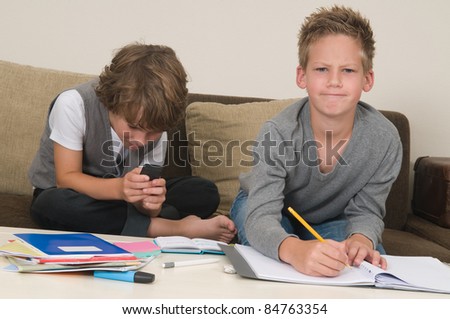 Two friends doing their homework in the livingroom. One of them is gaming, the other one is not ready and is jealous.