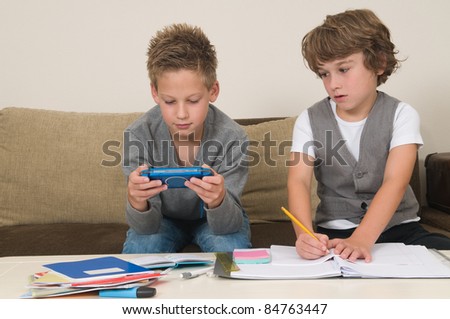 Two friends doing their homework in the livingroom. One of them is gaming, the other one is not ready and is jealous.