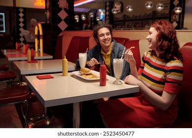 Two friends are enjoying milkshakes and burgers in a classic American diner with red seating booths, decorative elements, and soft lighting, creating a nostalgic atmosphere - Powered by Shutterstock