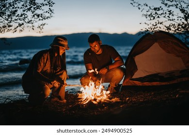 Two friends gather around a glowing campfire near a lake, preparing food as dusk settles, highlighting the beauty of the great outdoors. - Powered by Shutterstock