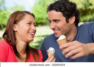 Two friends laughing while looking at each other and holding ice cream in a sunny park