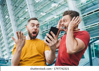Two friends looking extremely excited getting good news about winning a bet in online bookmaker watching broadcast with winner results on mobile phone. Football stadium on the background. - Shutterstock ID 1797742321