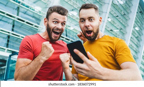 Two friends looking extremely excited getting good news about winning a bet in online bookmaker watching broadcast with winner results on mobile phone. Football stadium on the background. - Shutterstock ID 1836072991
