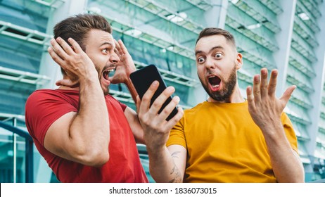 Two friends looking extremely excited getting good news about winning a bet in online bookmaker watching broadcast with winner results on mobile phone. Football stadium on the background. - Shutterstock ID 1836073015