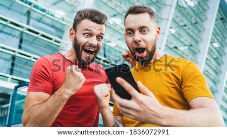 Two friends looking extremely excited getting good news about winning a bet in online bookmaker watching broadcast with winner results on mobile phone. Football stadium on the background.