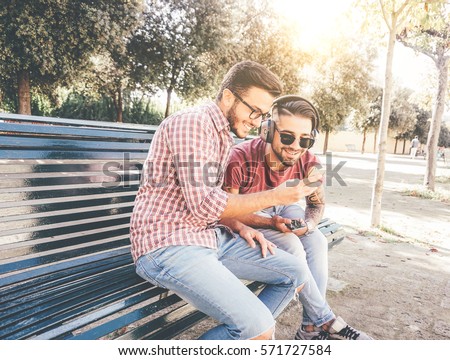 Two friends mates sitting on a bench in a park on a sunny day watching funny videos on social media with mobile smart phone - Two happy young men standing outdoor listening to music relaxing on bench