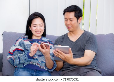 Two Friends On Line With Multiple Devices And Talking Sitting On A Sofa In The Living Room In A House Interior, Communication Concept.