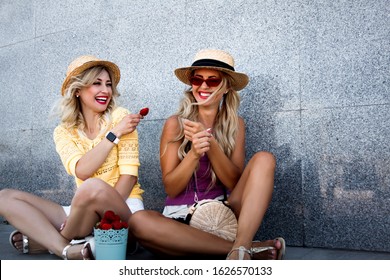 Two friends prefer healthy fresh fruit food in summer time. Women holding sweet organic strawberry and eating snack. People posing at the background street grey wall, wearing stylish urban clothes 