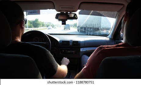 Two Friends Rides By Car On Urban Road. Men Traveling By Automobile At Summer Evening. Young Man In Sunglasses Driving Auto. View From Inside Of Car. Back View Close Up Slow Motion.