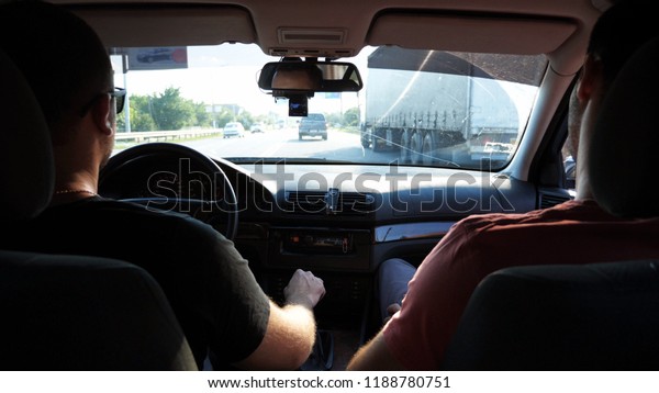 Two\
friends rides by car on urban road. Men traveling by automobile at\
summer evening. Young man in sunglasses driving auto. View from\
inside of car. Back view Close up Slow\
motion.